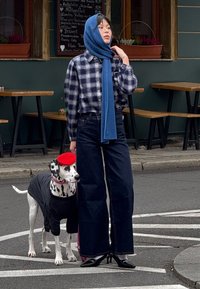 Modelo com camisa xadrez, cachecol azul e jeans largura larga escuros está ao lado de um cão vestido com boina vermelha e camisa preta, numa rua.