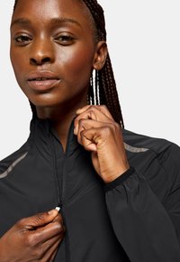 Chaqueta negra ligera con cremallera, que cuenta con puños elásticos y detalles reflectantes. El modelo está ajustando la cremallera en el cuello.