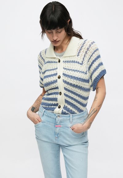 Πλεκτή ζακέτα με κοντά μανίκια σε κρέμ, ναυτικό και ανοιχτό μπλε ρίγες, με ριμπ γιακά και κουμπιά, φορεμένη πάνω από ανοιχτό μπλε τζιν.