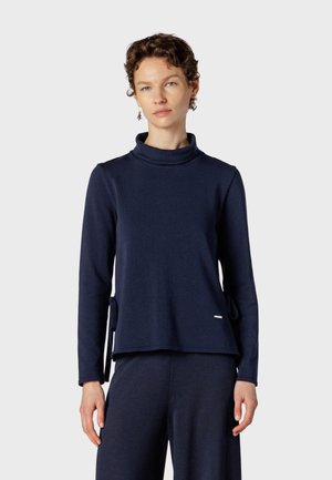 Femme aux cheveux courts et bouclés portant un haut à col roulé bleu foncé à manches longues et un pantalon large assorti sur un fond uni.