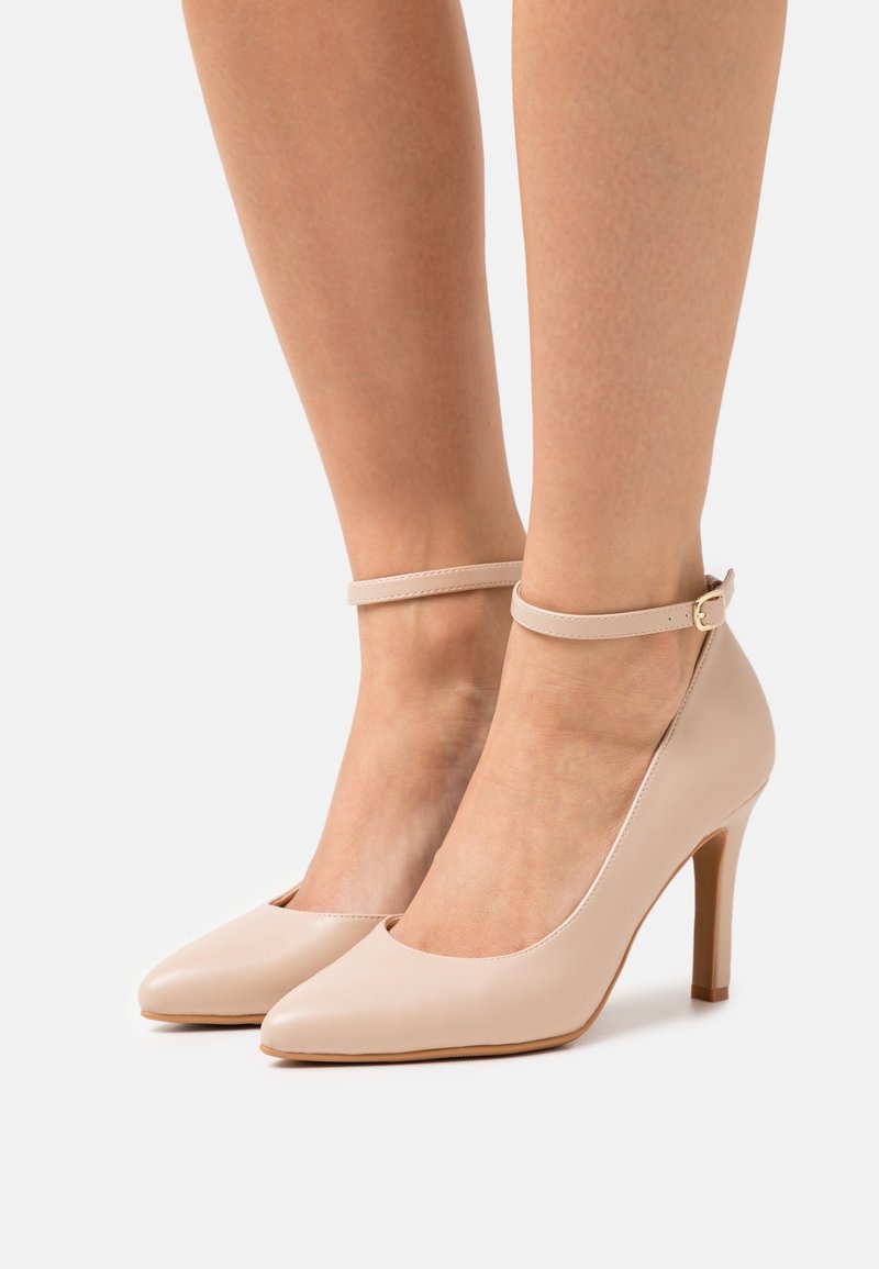 Pieds portant des talons hauts beiges à bout pointu avec des brides à la cheville et de petites boucles dorées sur fond blanc.