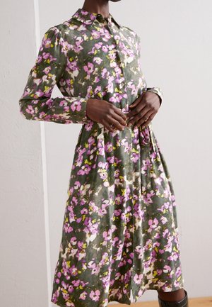 Kobieta ubrana w sukienkę z długim rękawem zapinaną na guziki, w kwiatuszki w kolorach różu, bieli i żółci na oliwkowozielonym tle.