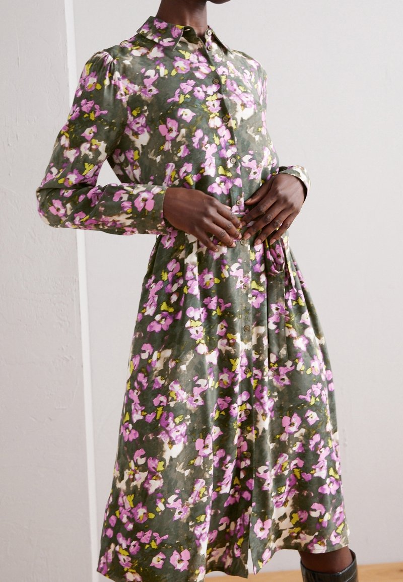 Femme portant une robe à manches longues boutonnée, à fleurs roses, blanches et jaunes sur un fond vert olive.