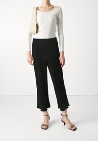 Femme portant un top blanc à épaules dénudées, un pantalon noir large et court, un sac à épaule beige et des sandales à talons en motif serpent.
