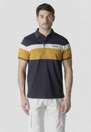 Hombre de pie mirando hacia adelante, vestido con una camiseta tipo polo de bloques de color azul marino, blanco y mostaza, y pantalones cortos cargo blancos, con un fondo liso.