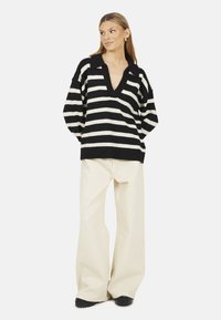 Pull rayé noir et blanc oversized avec un col en V et des manches courtes, associé à un pantalon large crème et des chaussures noires.