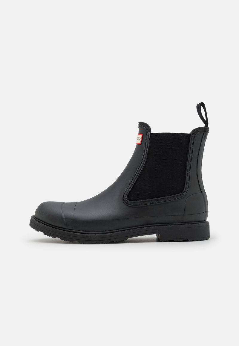 Bottes Chelsea en caoutchouc noir avec des panneaux élastiques sur les côtés, un bout arrondi et une semelle texturée. Comprend une languette à l'arrière pour un enfilage facile.