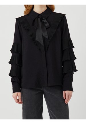 Czarna bluzka z satynową kokardą przy kołnierzu, falbanką w kształcie litery V na przodzie oraz warstwowymi plisowanymi falbanami na rękawach, noszona z wyblakłymi czarnymi jeansami.