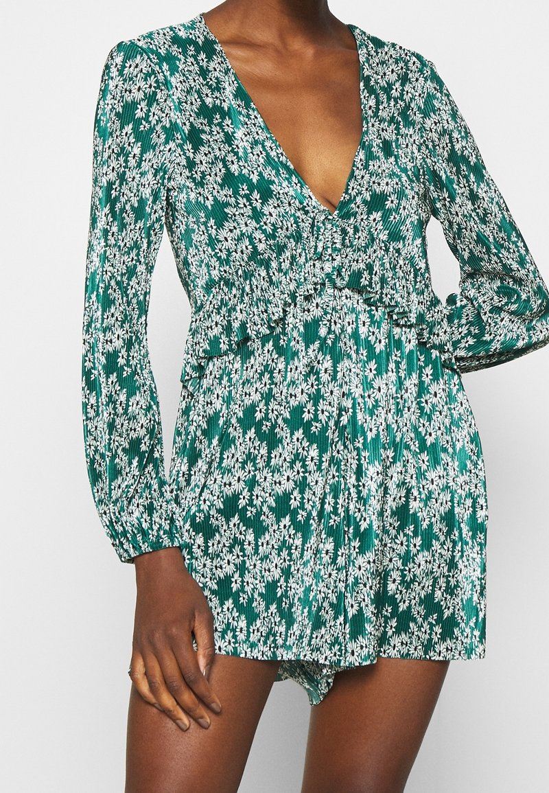 Femme portant une robe verte à manches longues avec un imprimé floral de marguerites blanches et un profond décolleté en V.