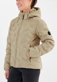 Beige quiltad jacka med huva, som har en frontdragkedja, sidofickor och en rund applikation på vänster ärm. Slät, lättviktsmaterial.
