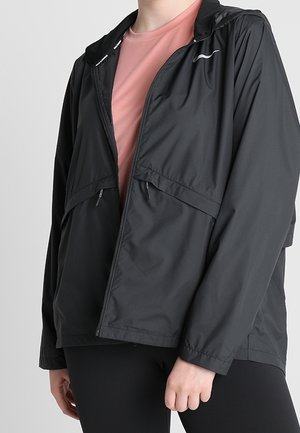 Person wearing black zip-up hooded jacket over pink shirt and black pants, shown from neck to hips, standing against light background.