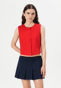 Top rojo corto sin mangas con un escote redondo y seis botones frontales, combinado con una falda corta plisada azul marino. Acabado de tejido texturizado.