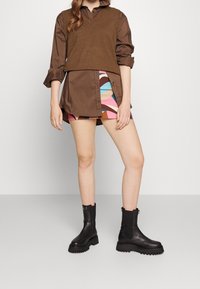 Chemise marron à manches longues superposée sous un gilet marron, associée à une jupe à motifs géométriques multicolores et des bottines noires à semelles épaisses.