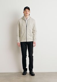Felpa zip-up beige chiaro, pantaloni neri, cappellino nero, maglietta bianca. La felpa presenta una tasca frontale e un cordoncino, con una texture di tessuto morbido.