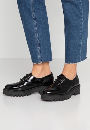 Person iført stramme blå jeans og sorte skinnende læder snørestøvler med tykke rillede såler, stående på et lyst gulv.