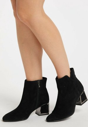 Beine, die schwarze Wildleder-Stiefeletten mit seitlichem Reißverschluss und Blockabsätzen tragen und auf einer weißen Oberfläche stehen.