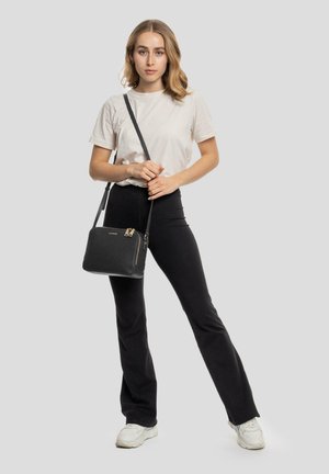 Sac à bandoulière en cuir noir avec des accessoires en métal doré, de forme rectangulaire, surface texturée et fermeture zippée. Modèle portant une tenue décontractée.