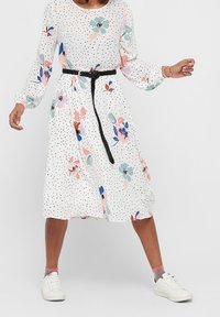 Nainen, jolla on päällään valkoinen, pitkähihainen midimekko, jossa on mustia pisteitä ja värikkäitä kukkakuoseja, musta letitetty vyö, harmaat ja vaaleanpunaiset sukat sekä valkoiset tennarit.