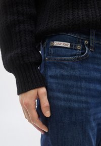 Μαύρο πλεκτό πουλόβερ με ριμπ υφή· μπλε τζιν παντελόνι με ετικέτα Calvin Klein, ασημένια κουμπιά και κλασικό σχέδιο με πέντε τσέπες.