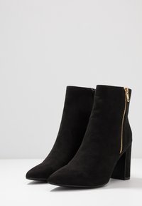 Botas de tobillo de ante negro con punta afilada, cremallera lateral dorada y tacón grueso. Textura suave sin patrones visibles.