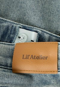Denim z jasnoniebieskim odcieniem, z brązową skórzaną metką wytłoczoną napisem "Lil'Atelier." Zawiera widoczne szwy i szczegół z guzikiem.