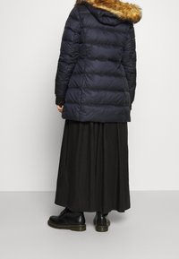Marineblauwe pufferjack met een gevoerde capuchon van bont, met horizontale stiksels en zijzakken, in combinatie met een zwarte lange rok en zwarte laarzen.