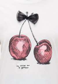 Camiseta blanca con un gráfico dibujado a mano de dos cerezas en rosa con contornos negros, adornado con un pequeño lazo negro de gasa encima.