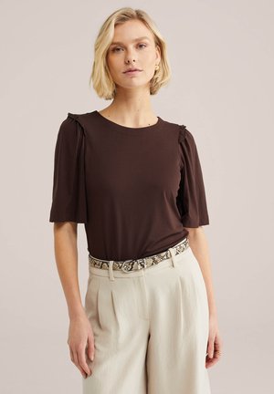 Lyshåret kvinde iført en mørkebrun kortærmet top med flæseærmer, lysebeige højtaljede bukser og et bælte med slangeskind.