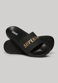 Sandales noires à glissière avec une large bande portant le texte doré "SUPERDRY". Semelle extérieure noire texturée avec motif de marque. Fabriquées en matériau synthétique.
