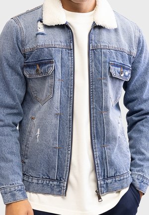 Mann trägt hellblaue Jeansjacke mit weißem Fleece-Kragen über weißem Hemd, Hände teilweise in den Taschen, vor einfarbigem Hintergrund.