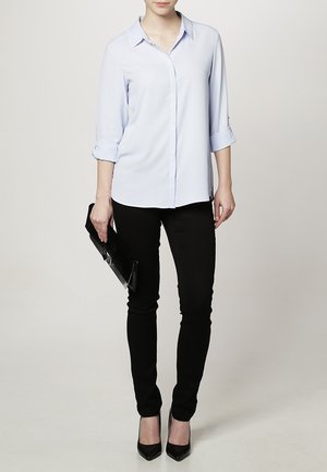 Camicia azzurro chiaro con maniche arrotolate, abbinata a jeans skinny neri e tacchi alti neri. La modella tiene una clutch nera.