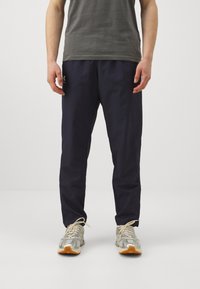 Pantalons de sport navy en tissu léger avec une taille élastique et des poches latérales, associés à un t-shirt gris et des chaussures de course argentées.