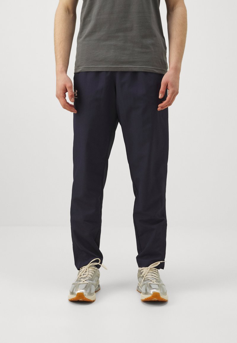 Pantalons de sport navy en tissu léger avec une taille élastique et des poches latérales, associés à un t-shirt gris et des chaussures de course argentées.