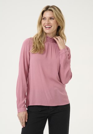 Blouse à manches longues, en tissu texturé de couleur rose clair, avec un col haut à volants et des poignets boutonnés. Portée avec un pantalon noir.