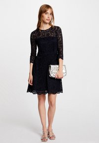 Robe en dentelle marine à manches trois-quarts, ourlet festonné et taille ajustée. Associée à une pochette en argent texturée et des sandales à lanières.