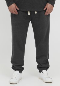 Un pantalon de survêtement gris foncé en tissu doux, avec une taille élastique à cordon de serrage et des chevilles resserrées. Porté avec des baskets blanches.