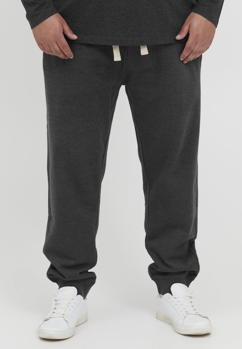 Un pantalon de survêtement gris foncé en tissu doux, avec une taille élastique à cordon de serrage et des chevilles resserrées. Porté avec des baskets blanches.