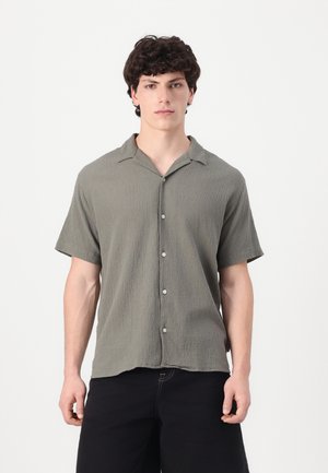 Short-sleeve, button-up shirt in textured, sage green fabric. Features a camp collar and subtle front pocket. Black shorts complete the look.