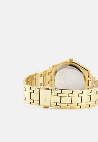 Montre en métal doré avec un bracelet à maillons, boîtier octogonal et dos en acier inoxydable visible. Présente une finition polie et un détail de logo.