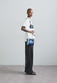 Geantă tote albastră cu "THE TOTE BAG" imprimat în alb, accesorii argintii, curea reglabilă, purtată cu pantaloni largi negri și un top alb.