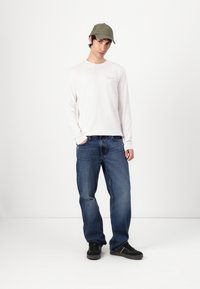 Wit T-shirt met lange mouwen en logo, blauwe spijkerbroek en olijfgroene pet. Model draagt zwarte sneakers en staat tegen een witte achtergrond.