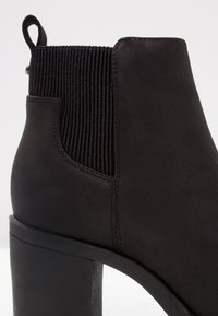 Stivale alla caviglia nero con una finitura in suede liscia, pannello laterale elastico testurizzato e tacco spesso. Presenta una linguetta di estrazione sul retro.