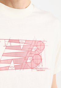 Camiseta de color crema claro con un diseño gráfico rojo que presenta formas geométricas y rayas, incluyendo el logo de "new balance" en la esquina inferior derecha.