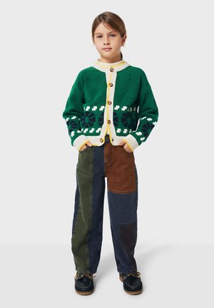 Jeune fille debout avec les mains dans les poches portant un cardigan à motifs verts et un pantalon en patchwork multicolore avec des chaussures foncées.