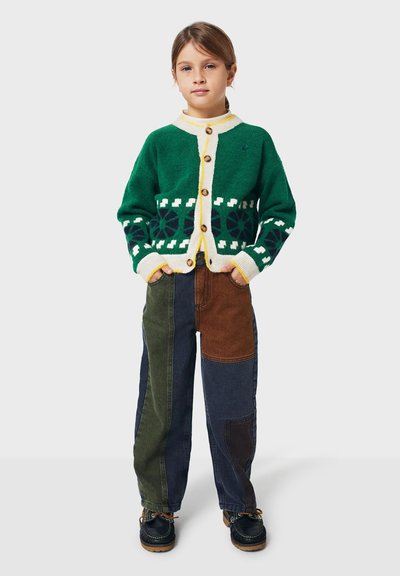 Ung flicka står med händerna i fickorna, klädd i en grönmönstrad kofta och flerfärgade lapptäckesbyxor med mörka skor.