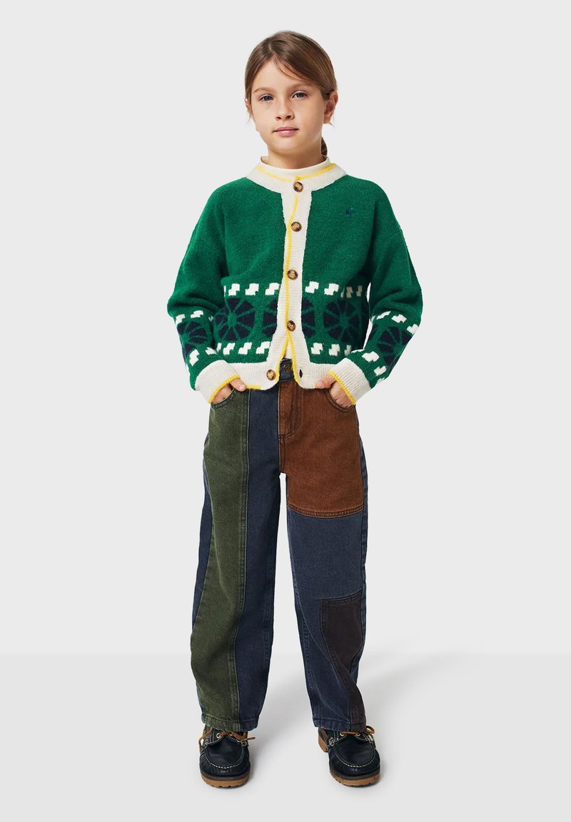 Junges Mädchen steht mit den Händen in den Taschen, trägt eine grün gemusterte Strickjacke und mehrfarbige Patchworkhosen mit dunklen Schuhen.
