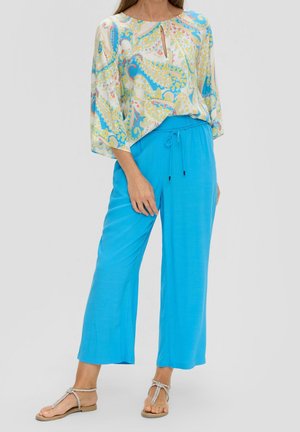 Kvinde iført en bluse med paisley-mønster og løstsiddende, lyseblå bukser med snøre, kombineret med sølvfarvede flade sandaler med stropper.