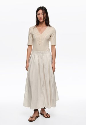 Kobieta stoi w beżowej bluzce z ażurowym wzorem i rękawami do łokcia, szerokiej, białej spódnicy w paski oraz brązowych sandałach typu thong.