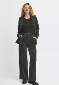 Črn suknjič s prugicami, ki je sestavljen iz krojenega jakne in širokih hlač; tkanina ima nežen sijaj; oblečen z črnim vrhom in čevlji.