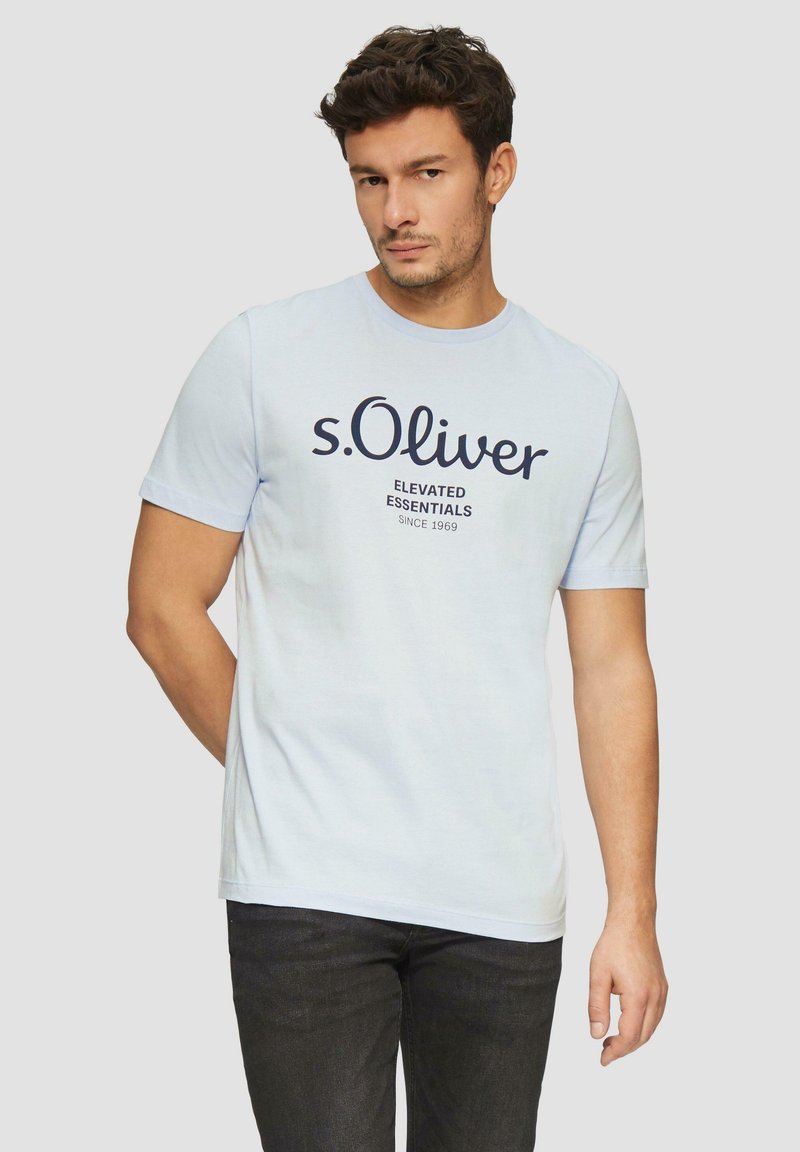 Heledsinine puuvillane T-särk lühikeste käiste ja ümarat kaelusega, millel on tume navy tekst: "s.Oliver Elevated Essentials alates 1969."